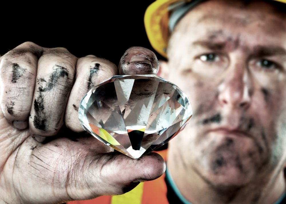 A diamond being held in a miners right hand with the camera focused on the diamond in the center of the image.